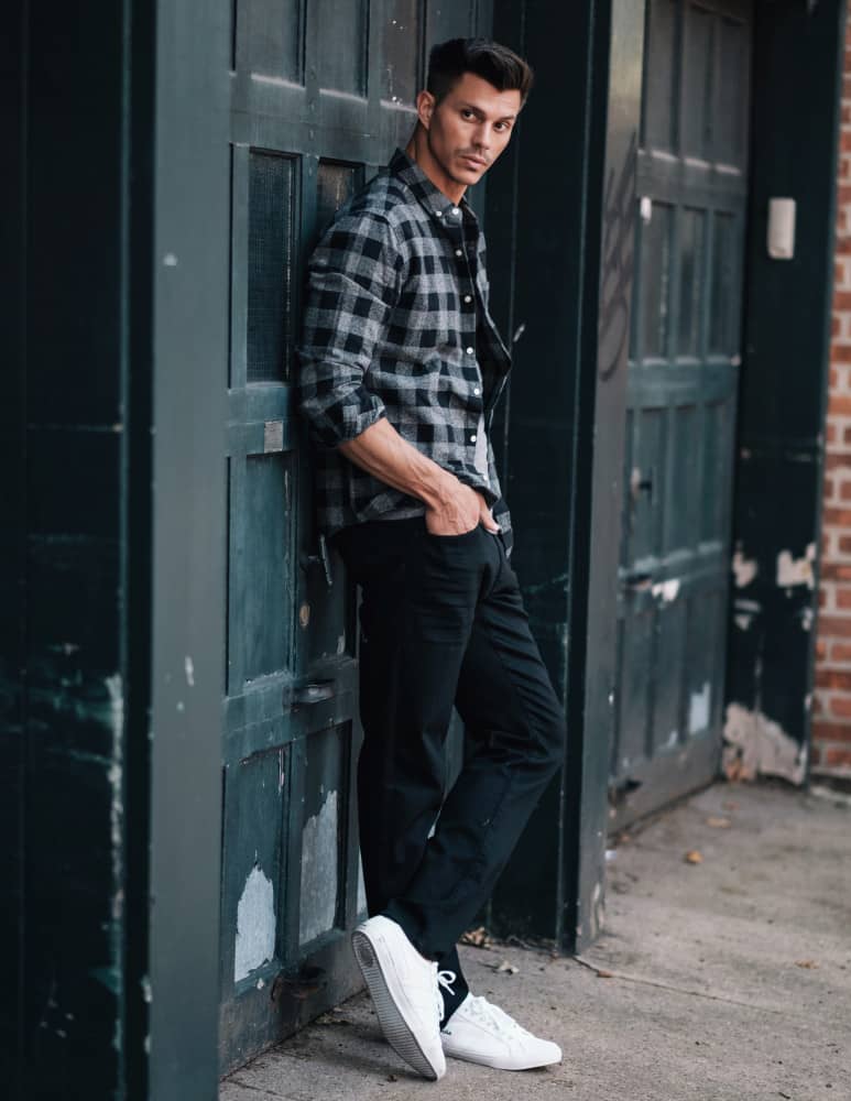 Kenny Braasch modeling a black and gray flannel shirt, black jeans, and white shoes while leaning against a garage door.